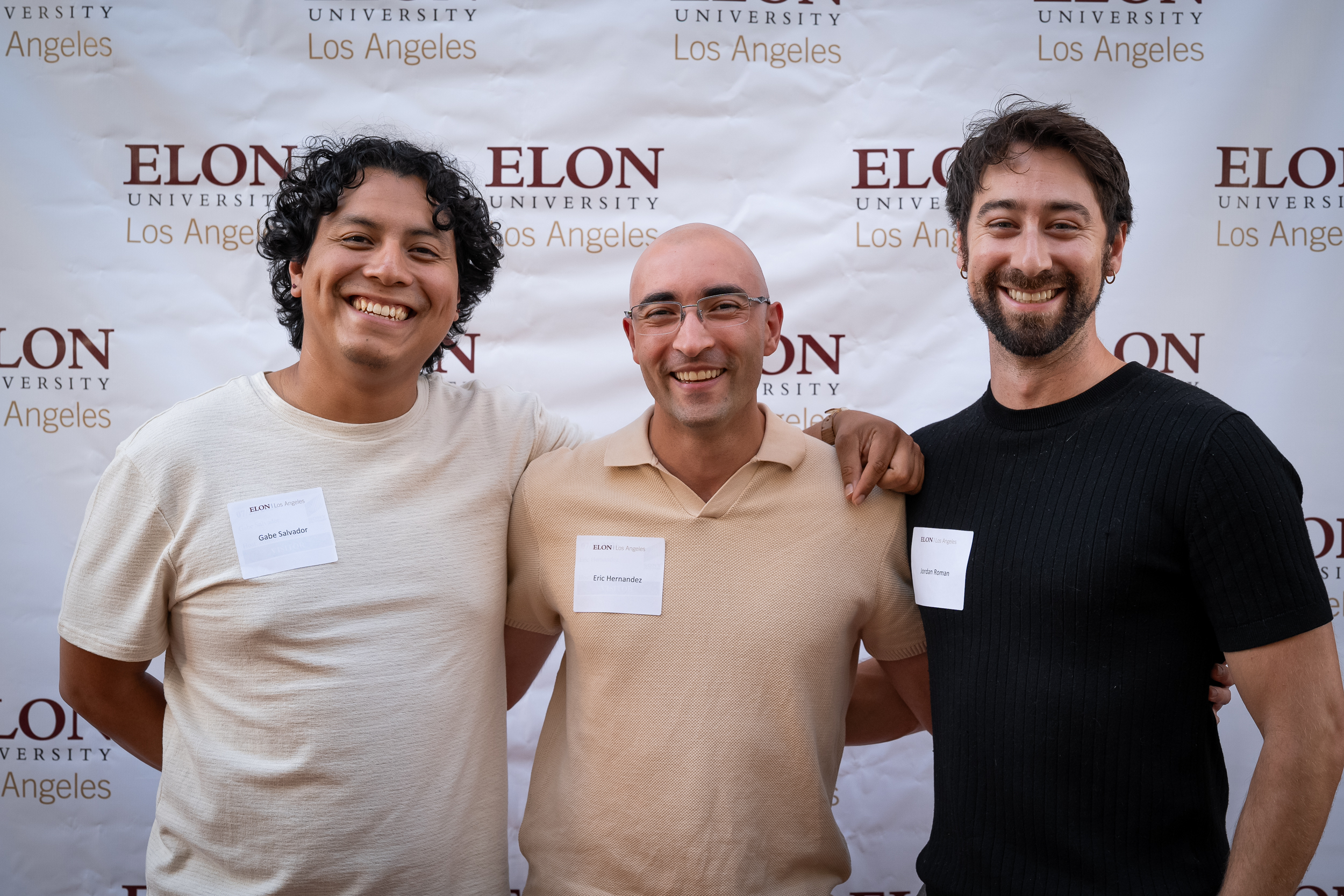 Three people pose for a photo in front of a white backdrop with Elon University Los Angeles logos