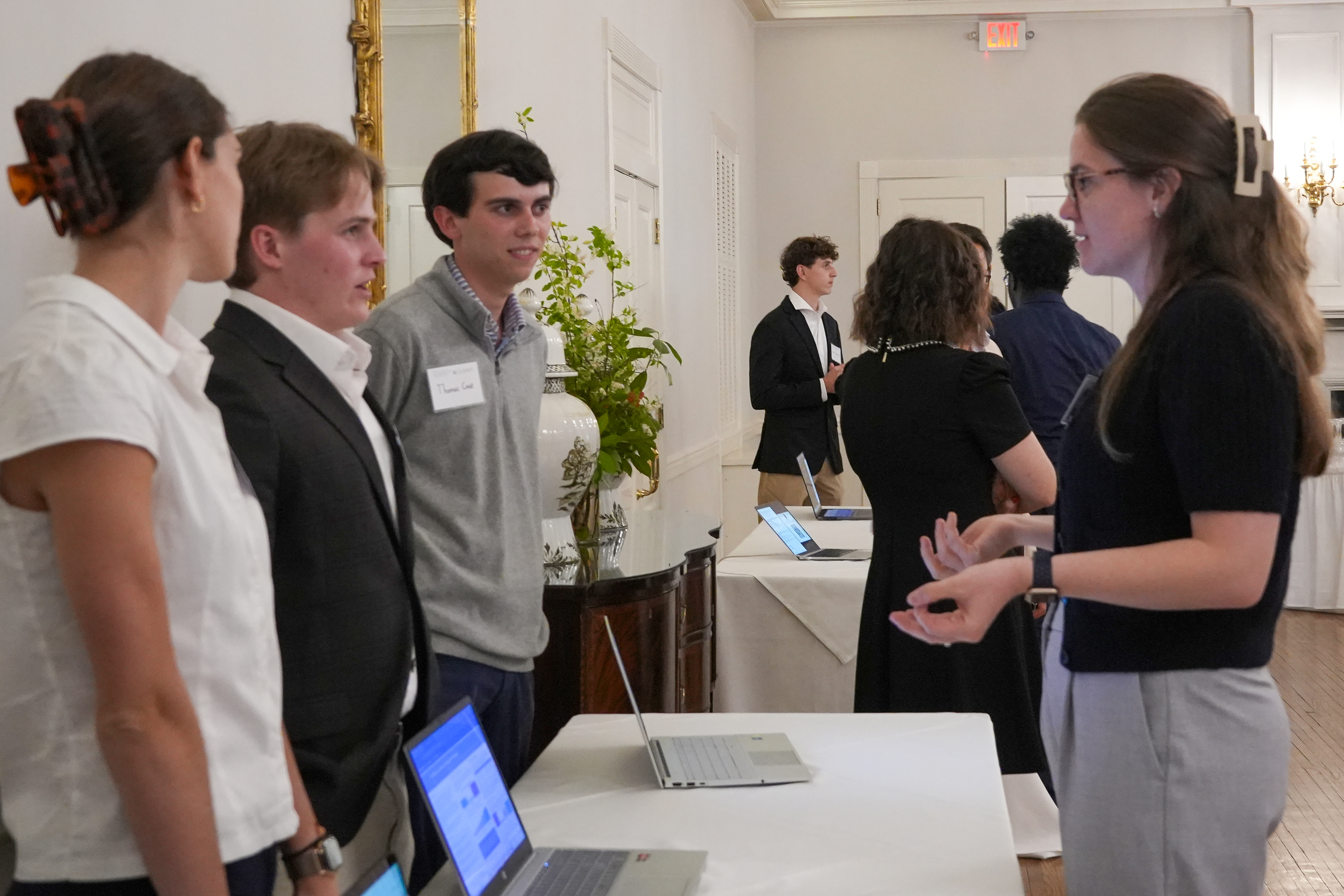 Thomas Case speaks with attendees during an AI showcase event. Laptops are open on tables as students discuss their projects.