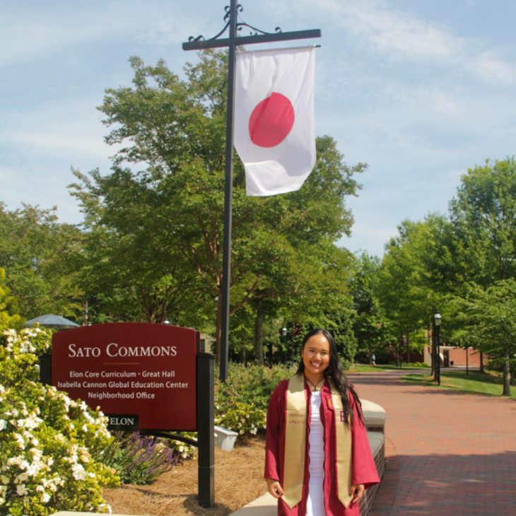 Sakura Kawakami stands near Sato Commons wearing Elon graduation regalia, with a Japanese flag displayed behind her.