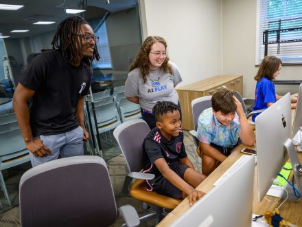 Man with black shirt and glasses and woman with glass and a tee shirt with the words AI Play peer over two boys sitting in front of computers looking at their screens.