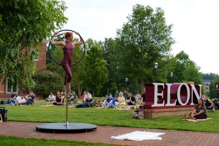 An aerial performer balances on a suspended hoop near the ���ϲ����� sign as community members relax on the lawn during an outdoor campus performance.