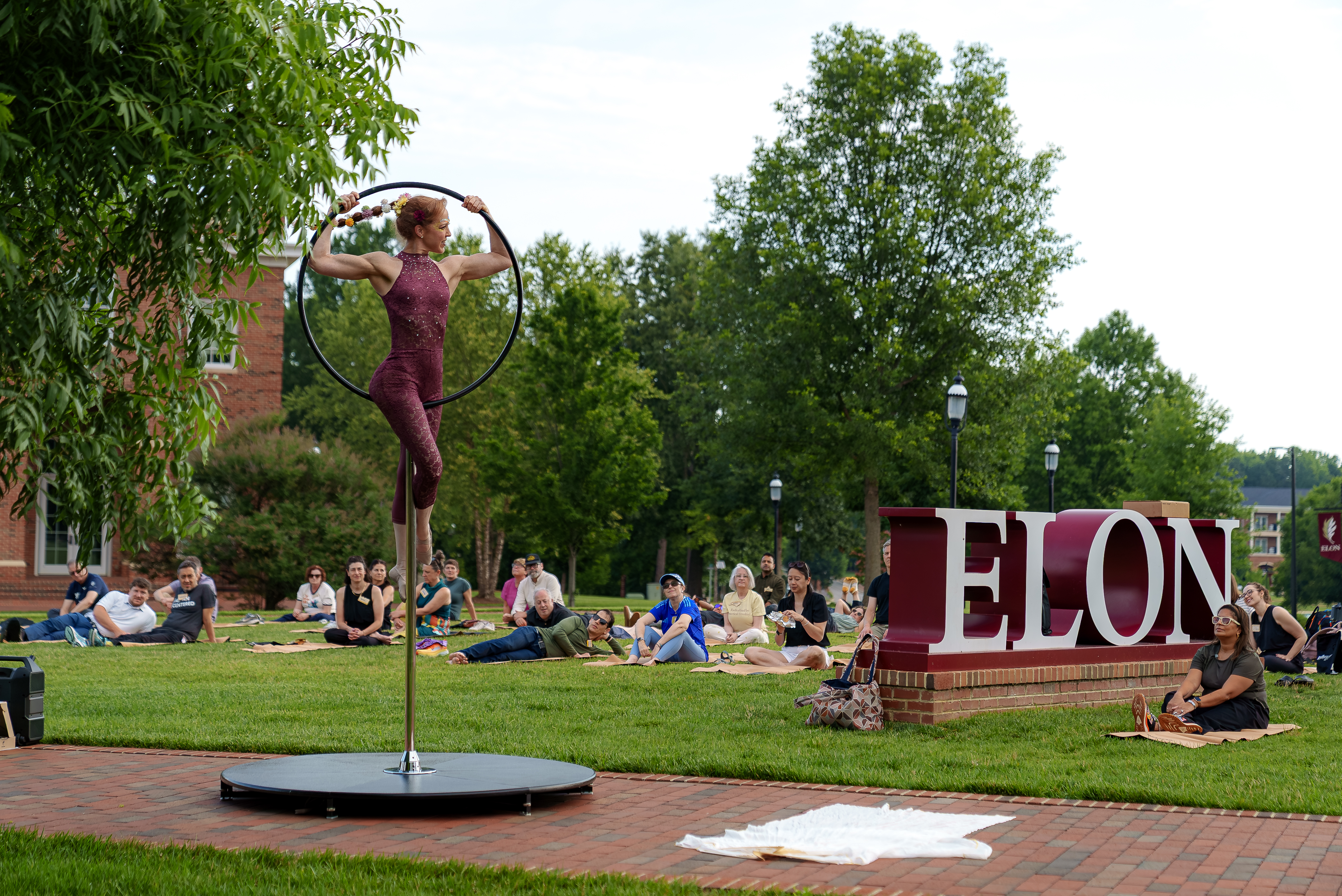 An aerial performer balances on a suspended hoop near the 51������ sign as community members relax on the lawn during an outdoor campus performance.