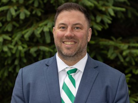 Professional portrait of a Ben Boivin in a gray suit and green and white diagnonally striped tie with lush green foliage in the background.