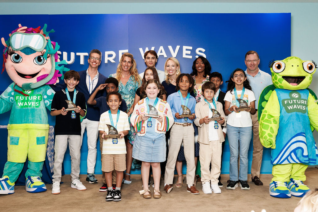 A group of children proudly display medals while posing with adult leaders and two colorful “Future Waves” mascots in front of a blue event backdrop.