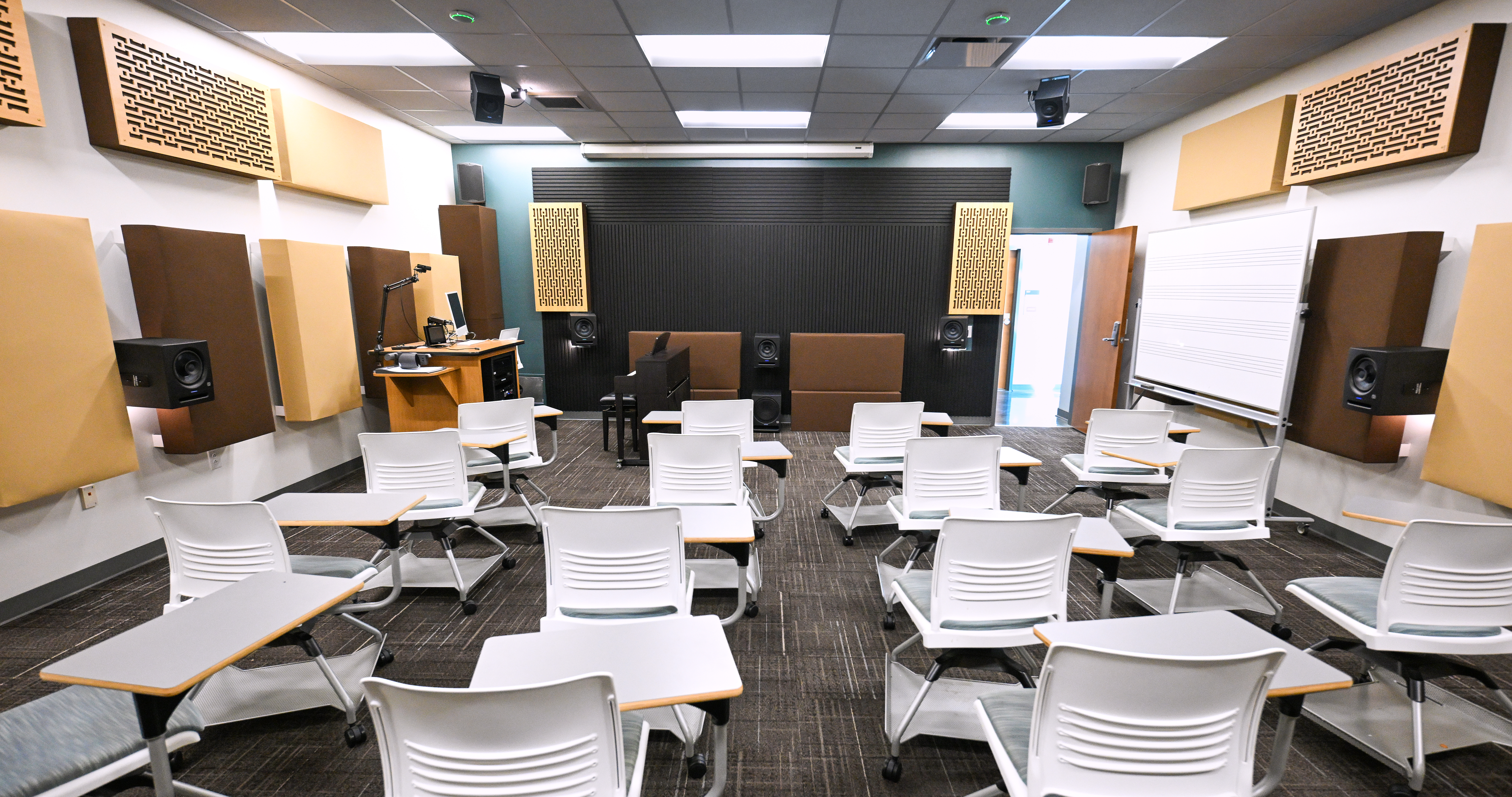 An empty audio production classroom features acoustic wall panels, studio monitors, a keyboard, instructor workstation, music staff whiteboard, and movable student desks arranged for instruction.