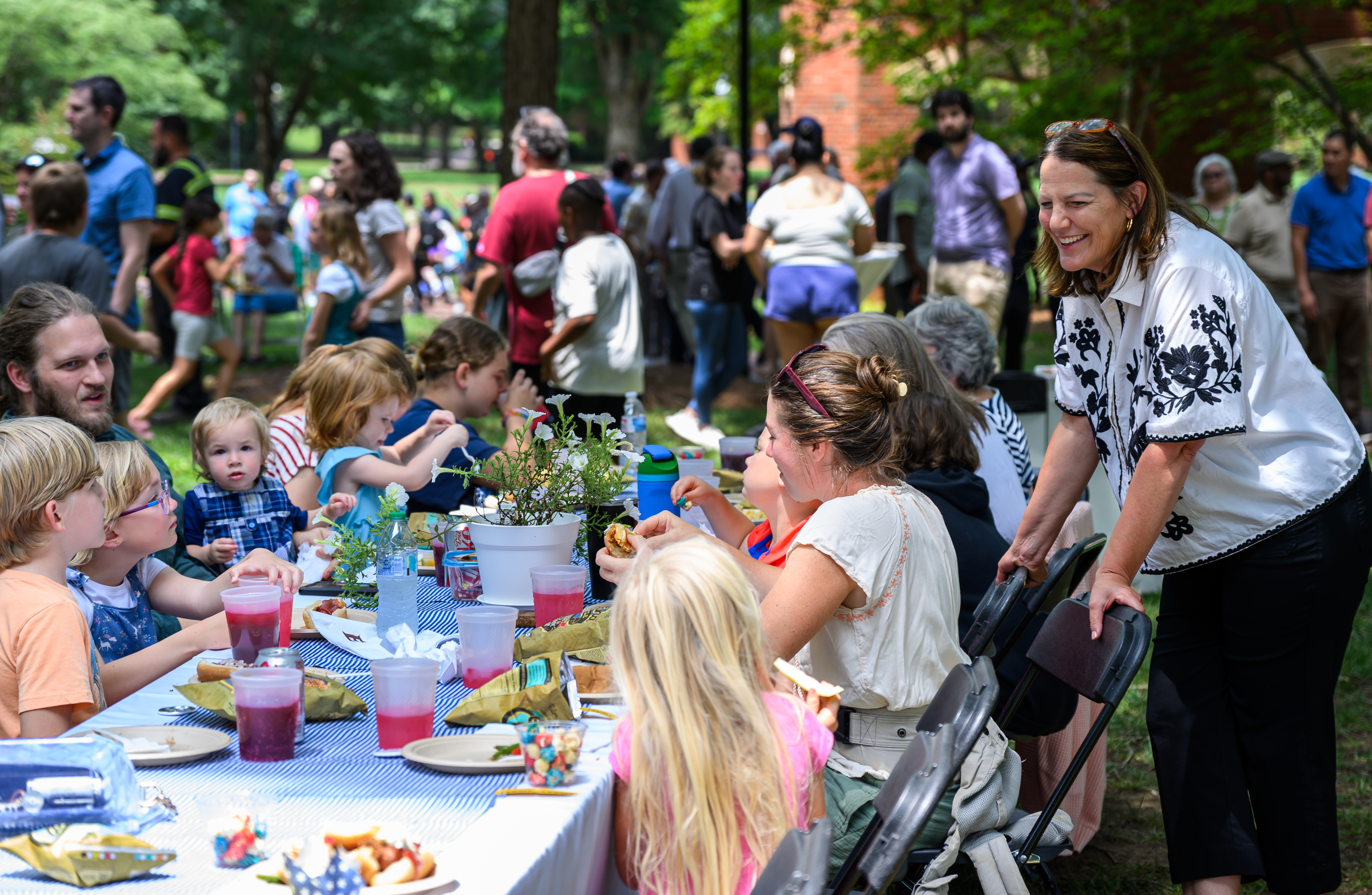 People of all ages gather around picnic tables on the university campus during a community event, sharing a meal and conversation outdoors while children and families participate in activities nearby.