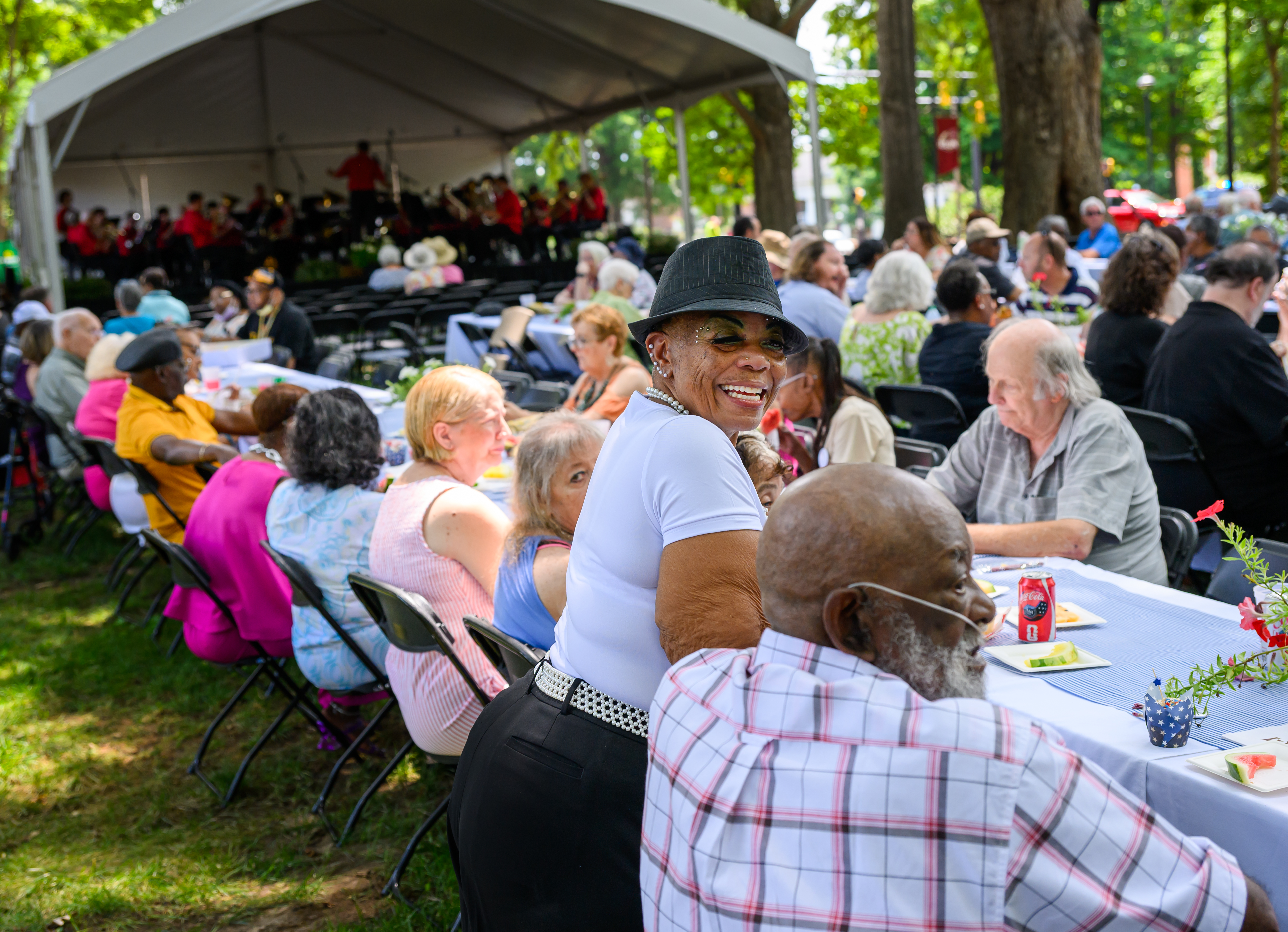 People sit around a long table during a community picnic. One woman in a hat and white shirt looks over her shoulder and smiles at the camera