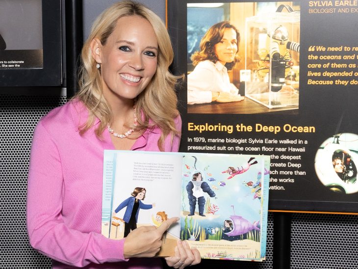 A woman in a pink shirt holds a book open pointing to a page while smiling