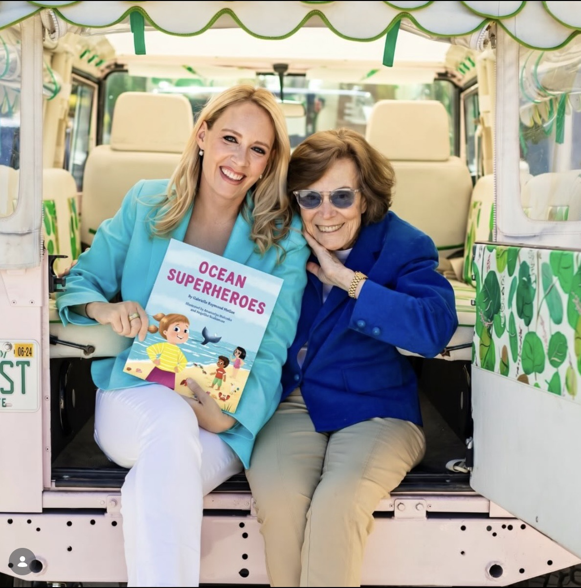 Two women sit side by side smiling for a photo. One holds a book called "Ocean Superheroes"