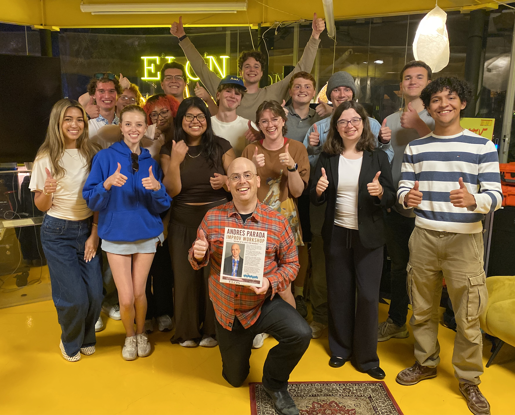 A group of students and a faculty member pose together in a media workspace, smiling and giving thumbs-up while one person holds a newspaper featuring the faculty member on the front page.