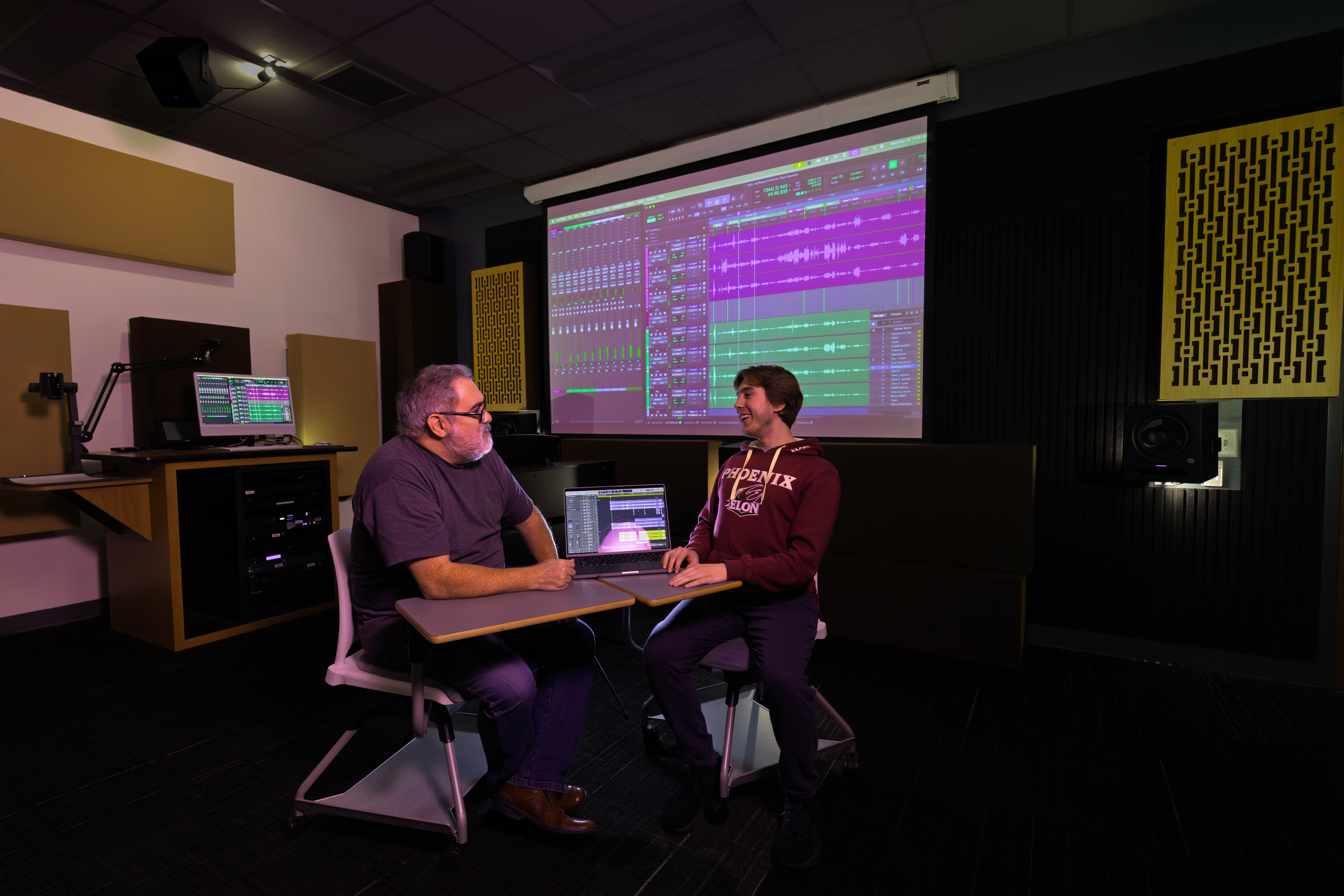A faculty member and student sit together in an audio production studio reviewing a multitrack recording project displayed on a laptop and large projection screen, discussing audio editing and sound production techniques.