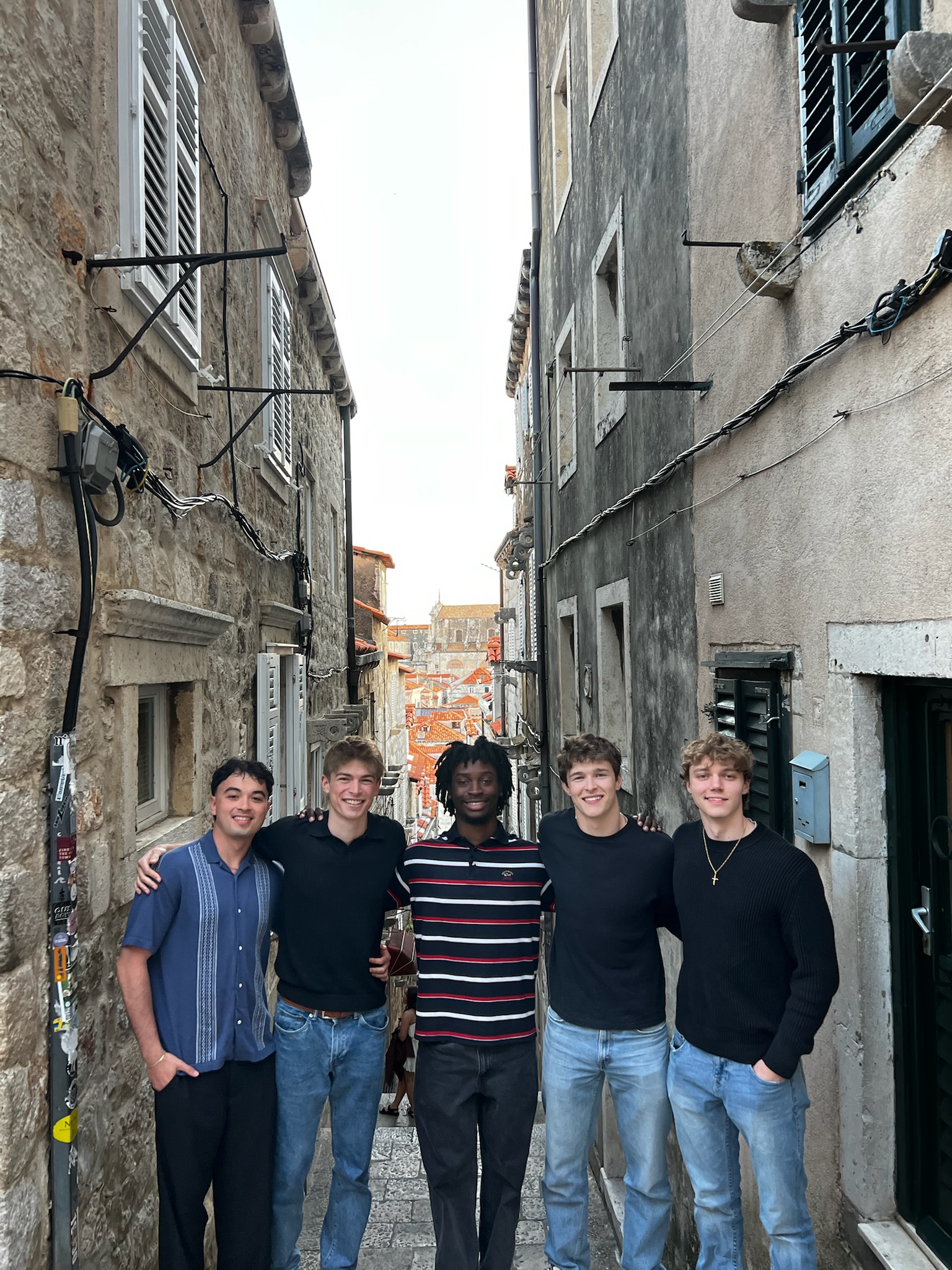 Five people stand arm in arm in a narrow stone alley lined with historic buildings and shuttered windows. The alley opens to a view of terracotta rooftops and a church tower in the distance, suggesting a European city setting.