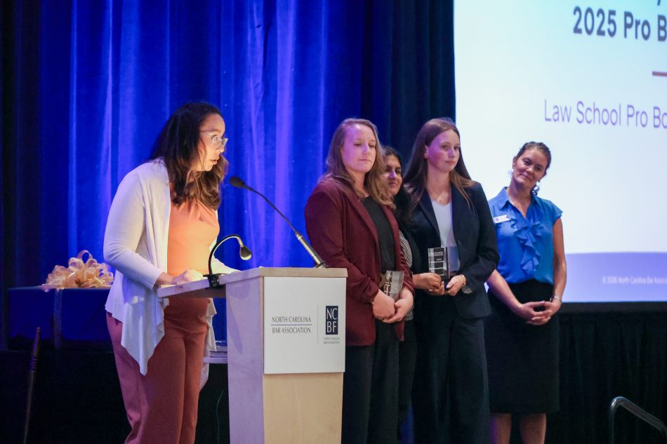 A person speaks into a microphone at a podium onstage as law students wait nearby to accept an award.