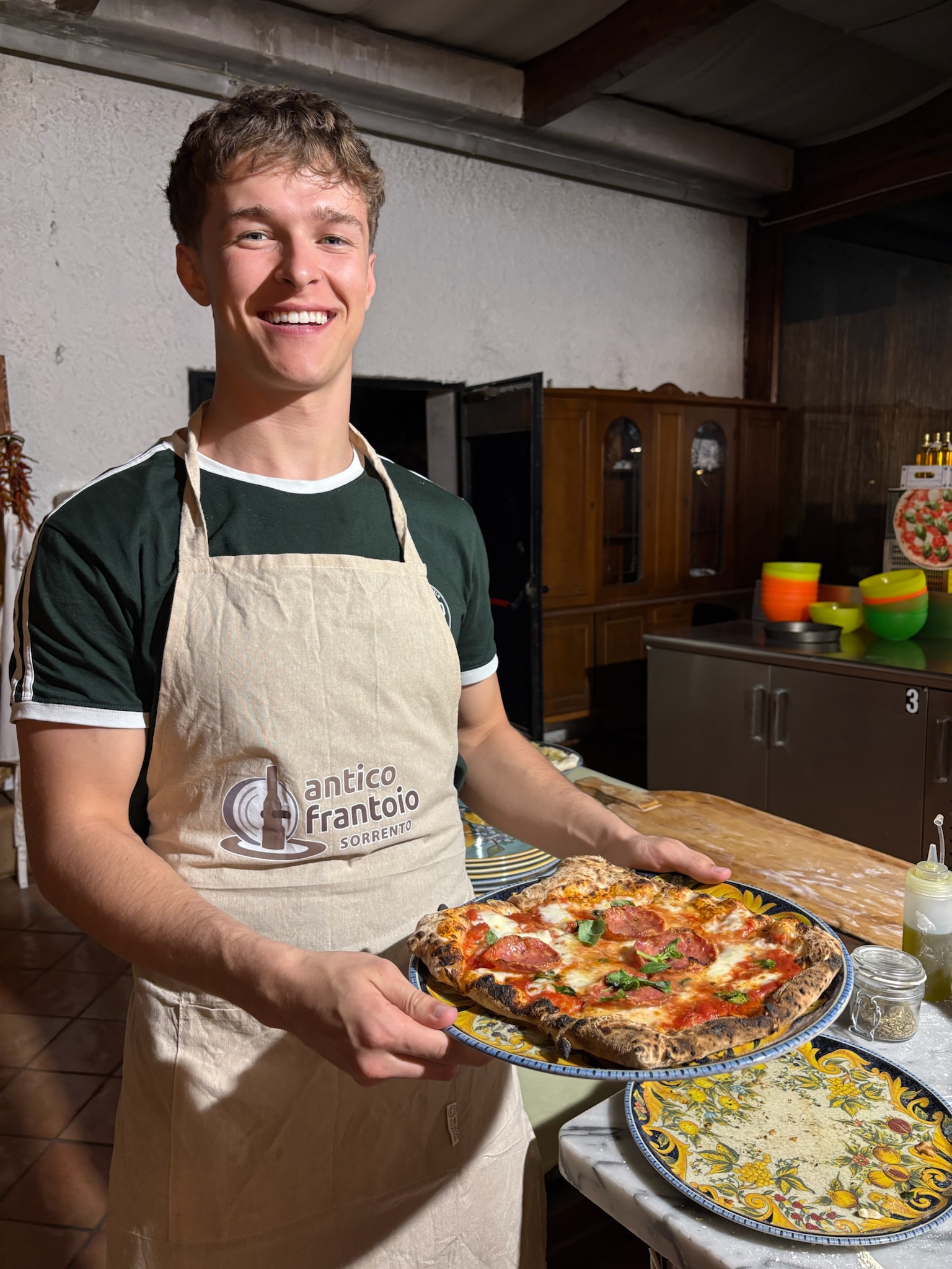 A smiling person wearing an apron labeled “Antico Frantoio Sorrento” holds a freshly baked pizza