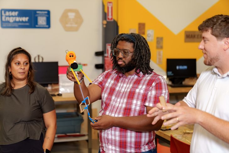 Three people stand in a makerspace as one participant demonstrates a 3D-printed mechanical device, explaining its design while others observe and discuss the project.