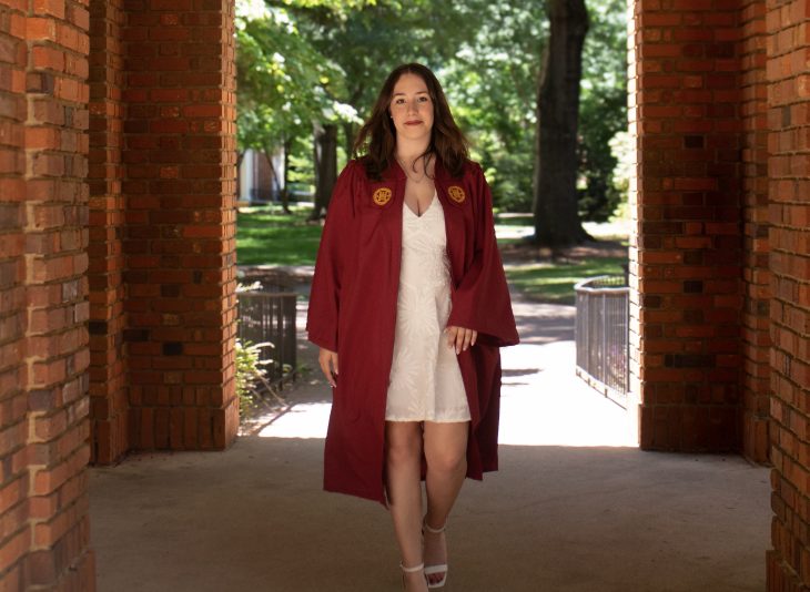 A woman in an open maroon graduation robe and a white dress walks through a brick archway