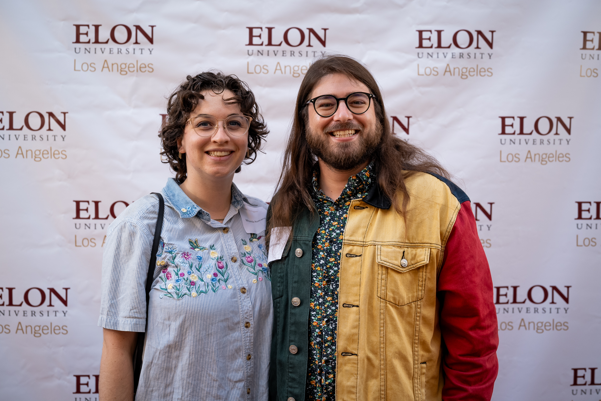 Two people pose for a photo in front of a white backdrop with the Elon University Los Angeles logos