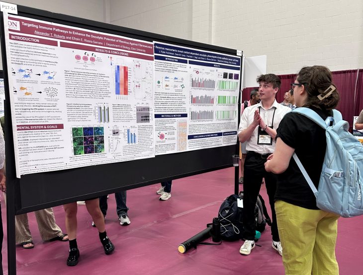  A student presenter stands beside two scientific research posters, explaining the findings to an attendee wearing a light blue backpack during a poster session at an academic conference.