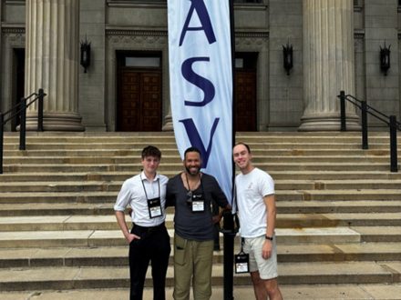 Three people pose for a photo in front of a building with two columns and steps. A tapestry that reads "ASV" is directly behind them.