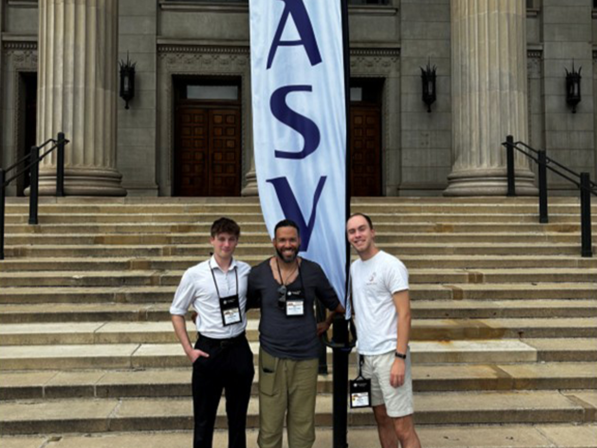 Three people pose for a photo in front of a building with two columns and steps. A tapestry that reads 