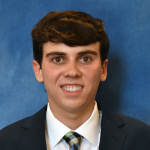 Thomas Case smiles for a headshot in a dark suit jacket, white shirt and patterned tie against a blue background.