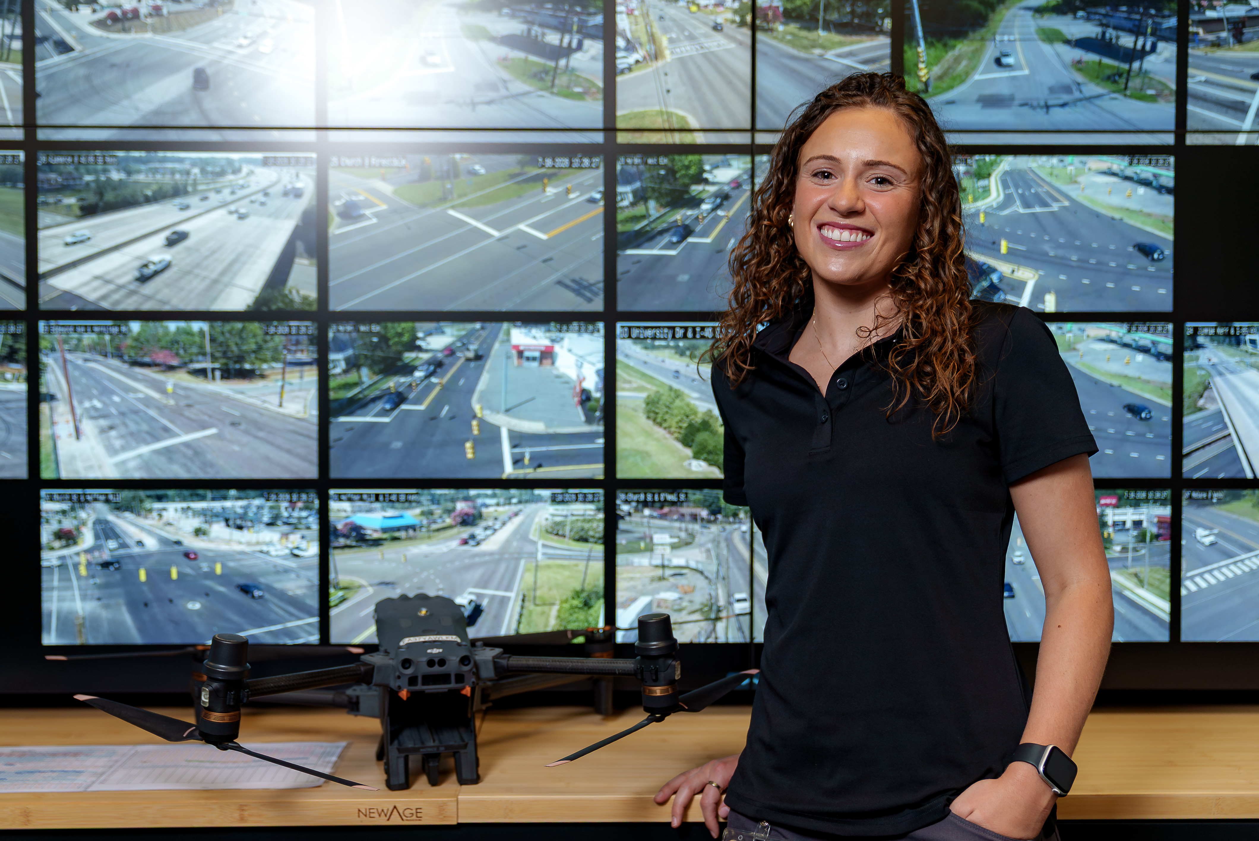A smiling person stands beside a drone in a traffic management or public safety operations center. Multiple large monitors behind them display live traffic camera feeds from intersections and roadways.