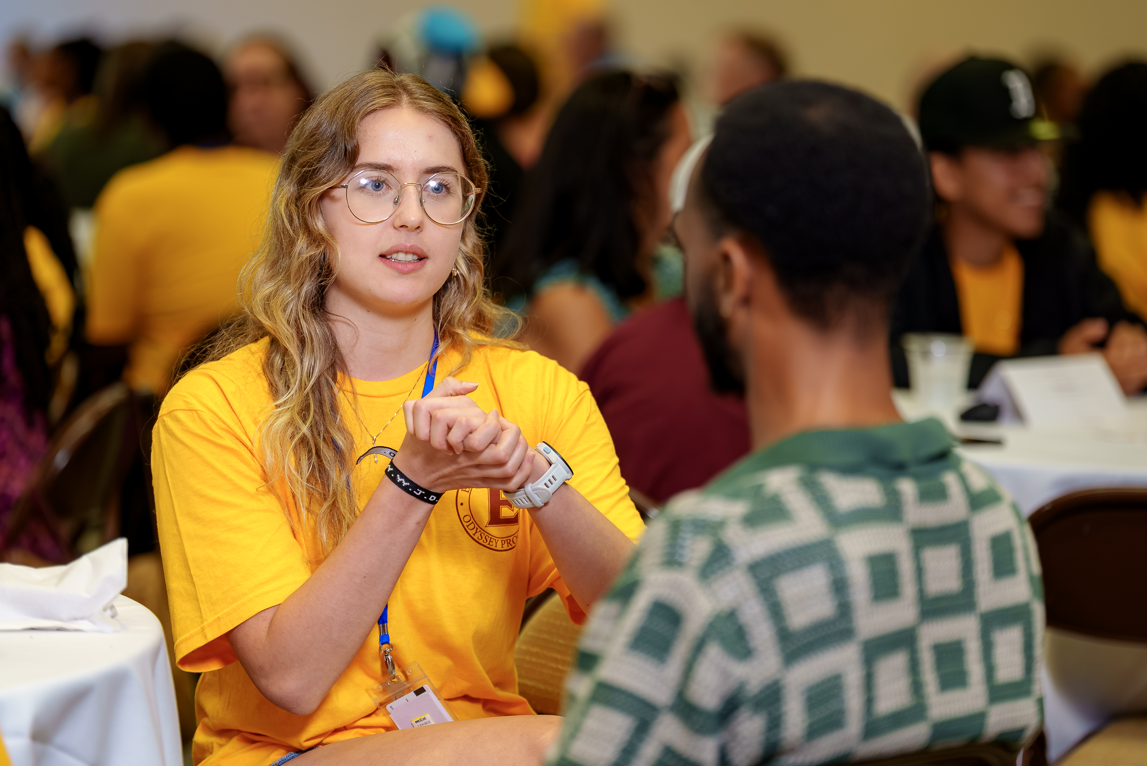 A student in a gold Odyssey Program T-shirt sits at a round table speaking with another attendee during a networking session. She gestures with her hands while talking, wearing glasses and a name badge, as other participants converse in the softly blurred background.
