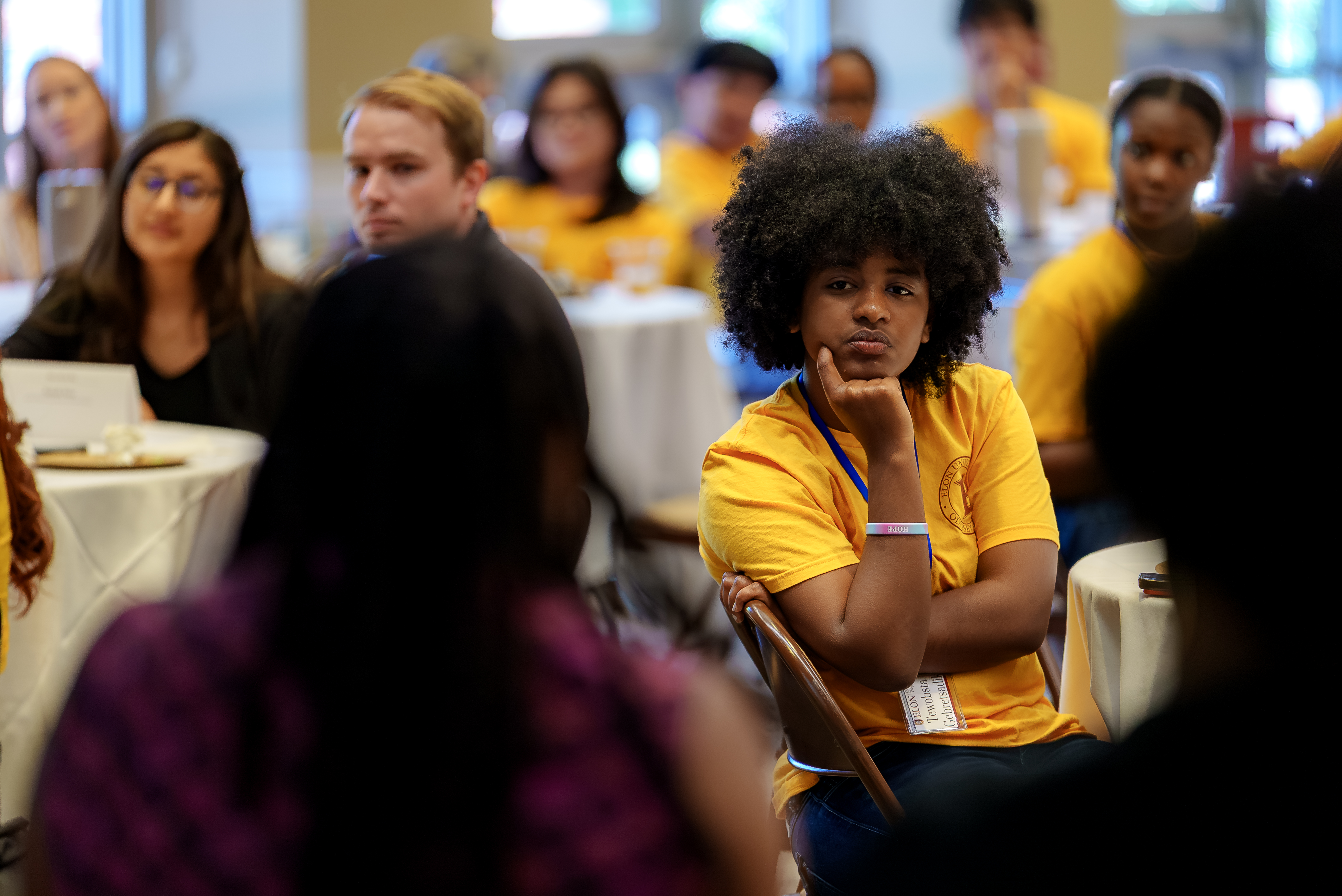 A student wearing a gold Odyssey Program T-shirt listens attentively during a group discussion, resting their chin on one hand while seated among peers at round tables. Other attendees are visible in the background, creating a thoughtful and engaged atmosphere.