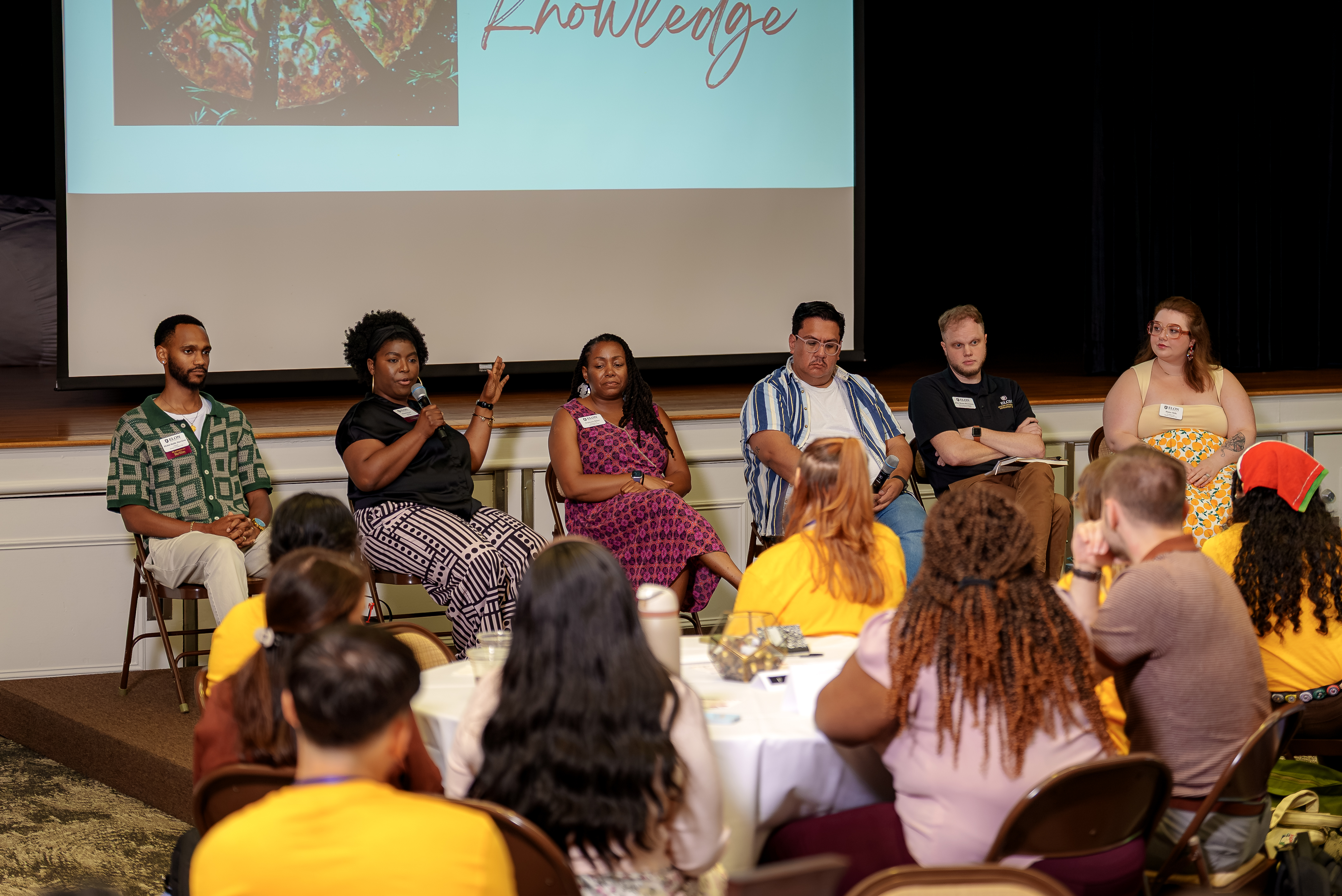 Six panelists sit across the front of a meeting room speaking with students gathered around round tables. One panelist gestures while speaking into a microphone as the others listen. A presentation slide projected behind them includes an image of pizza and the word “Knowledge,” reflecting the event’s discussion-focused atmosphere.