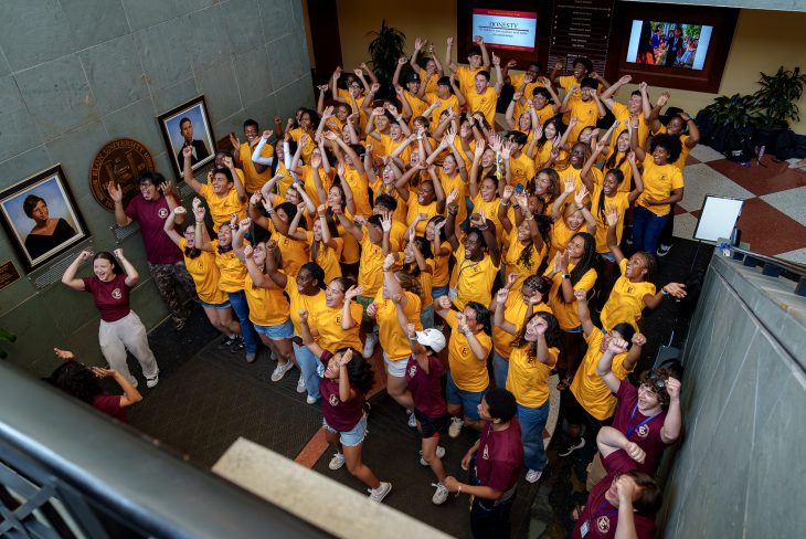 An overhead view shows dozens of students gathered in a campus atrium, many wearing bright gold T-shirts while others wear maroon shirts. The students cheer with their arms raised, smiling and looking upward toward the camera. Framed portraits and university signage line the walls, creating an energetic scene of celebration and community.