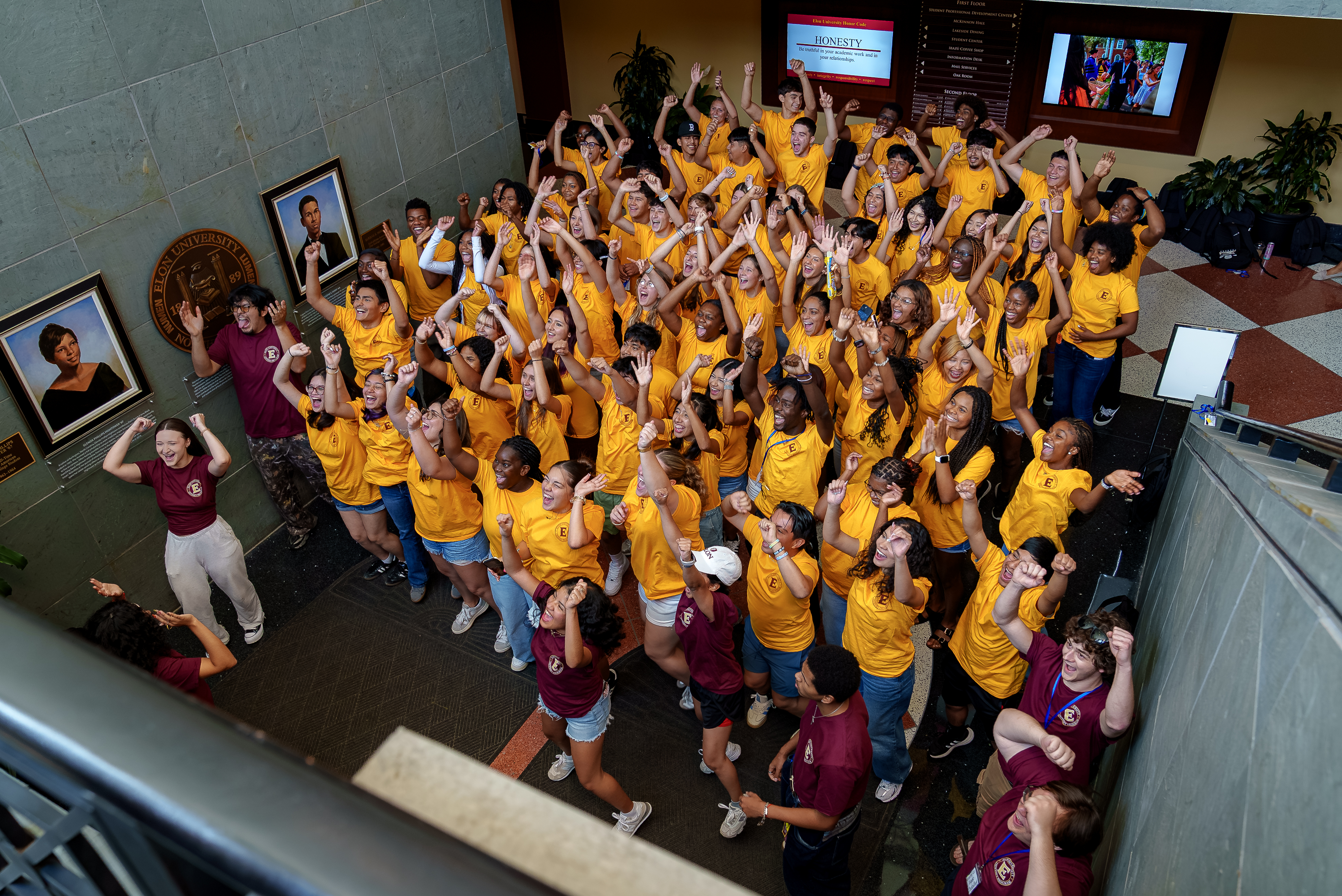 An overhead view shows dozens of students gathered in a campus atrium, many wearing bright gold T-shirts while others wear maroon shirts. The students cheer with their arms raised, smiling and looking upward toward the camera. Framed portraits and university signage line the walls, creating an energetic scene of celebration and community.