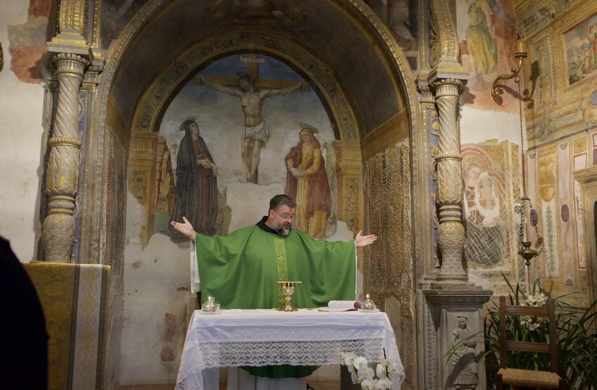 A priest wearing green liturgical vestments stands behind an altar with his arms outstretched during a church service. Behind him, a fresco depicting the crucifixion is framed by ornate columns and historic religious artwork covering the chapel walls.