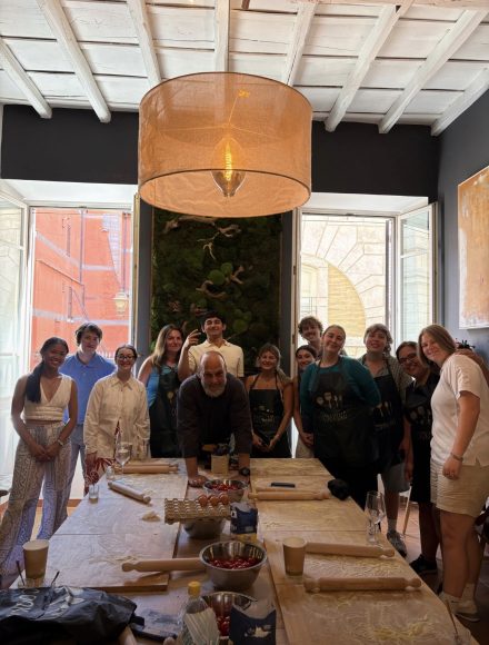 A group of students stands around a large worktable during a hands-on cooking class. Rolling pins, dough, and kitchen supplies are spread across the table, while large windows and contemporary artwork brighten the room behind them.