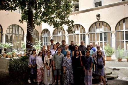 A group of approximately 20 people poses in a shaded courtyard surrounded by tall arched windows and potted plants. A large tree stands in the foreground, and the group is gathered on stone steps in the center of the historic-looking space.
