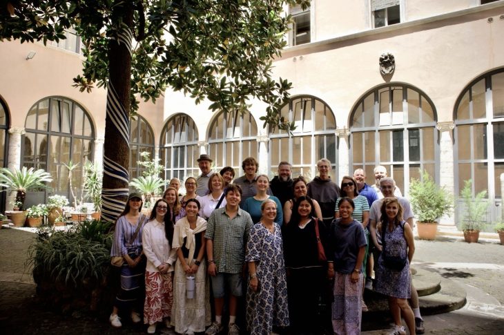A group of approximately 20 people poses in a shaded courtyard surrounded by tall arched windows and potted plants. A large tree stands in the foreground, and the group is gathered on stone steps in the center of the historic-looking space.