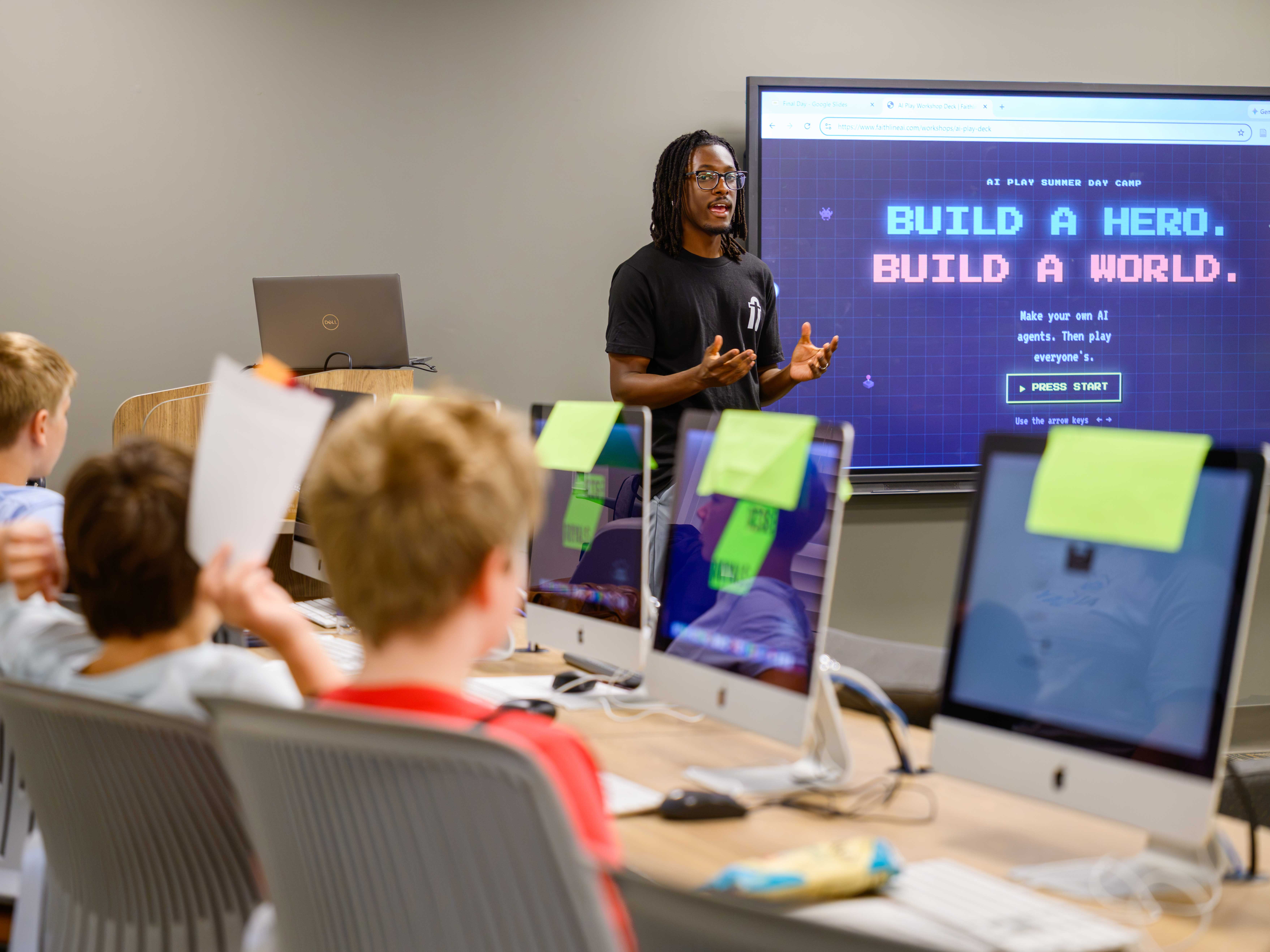 A man with a black tee shirt and glasses stands at the front of a classroom with a screen behind him that says "Build a Hero. Build a World." Middle school children are sitting at desks with computers looking up at the front of the classroom toward him.