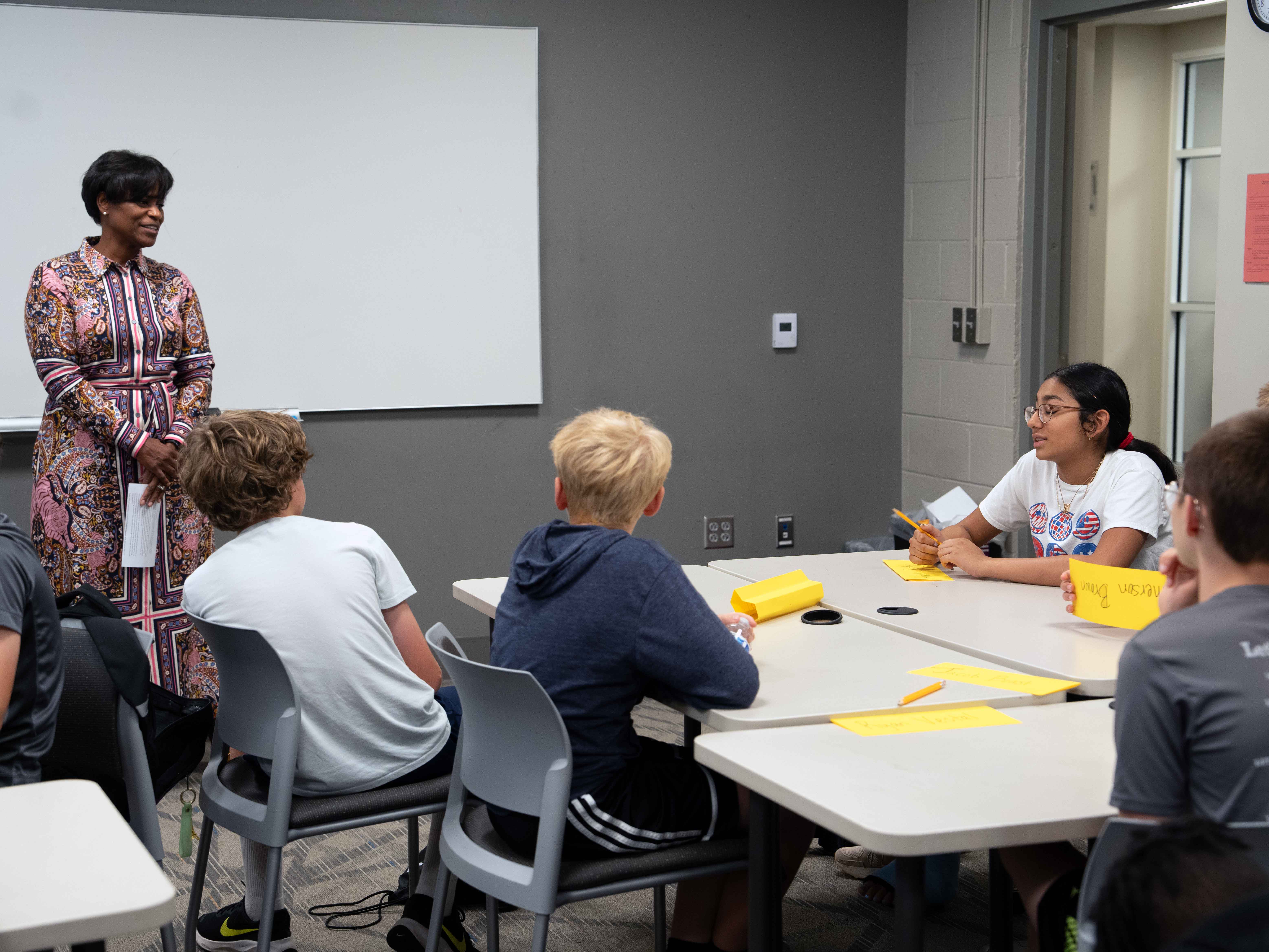 A woman in a floral dress stands in front of a white board in a classroom speaking with children sitting at desks.