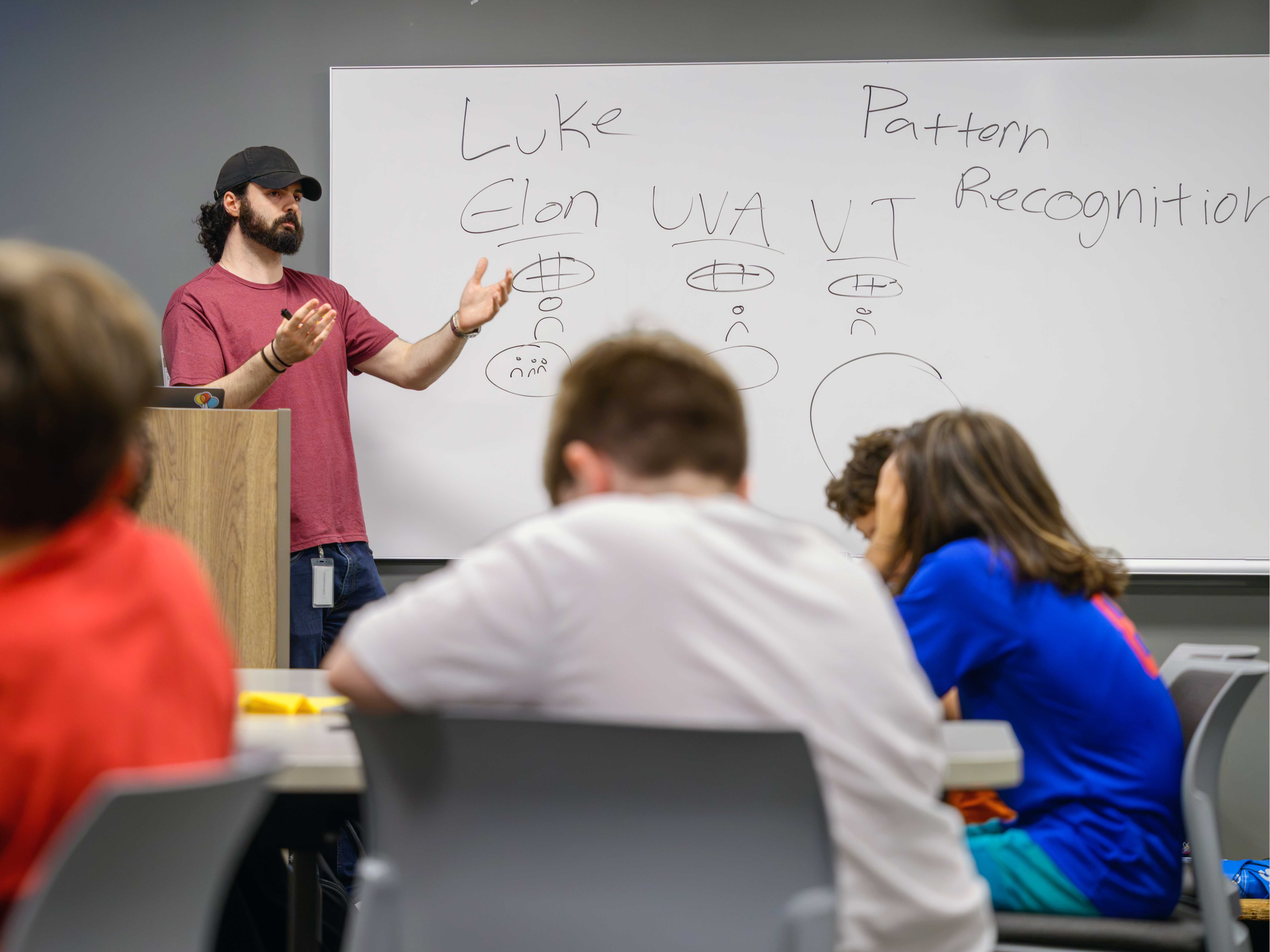 A man with a red tee shirt and baseball cap points to the word "Elon" on a white board in front of a classroom full of middle school children sitting at desks.