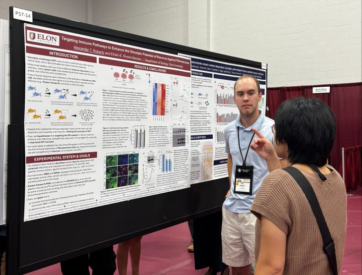 A student presenter stands beside a scientific research poster, listening as an attendee gestures with one hand while asking a question during a conference poster session.