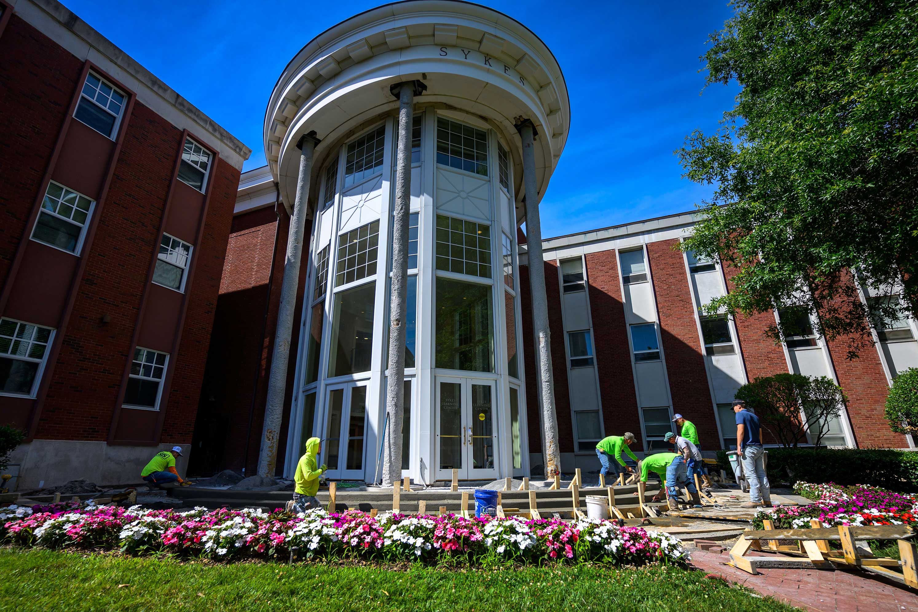 In the foreground, six construction workers repair the brick plaza in front of Sykes Learning Center. Pink and white flowers are planted along the perimiter of the walkway leading to the building.The exterior columns at the entrance of Syles Learning Center are are under renovation, exposing its bare foundation.