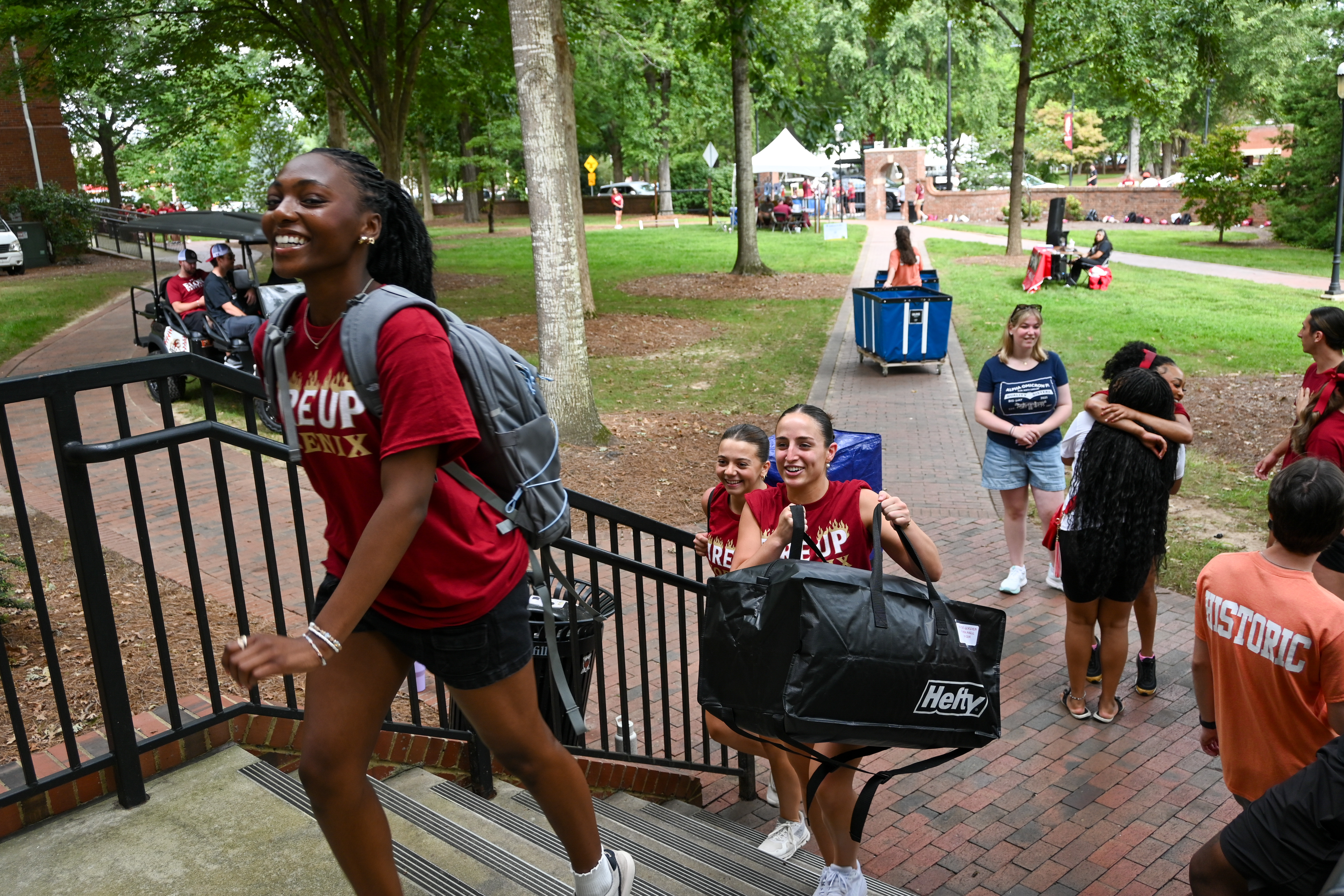 A group of people is moving up a staircase outdoors. One person is carrying a backpack, while two others are carrying a large bag labeled 