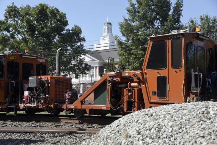 A photo of an orange Norfolk Southern rail maintenance piece of equipment on tracks near Elon University with Lindner Hall in the background and a pile of railroad ballast in the foreground.