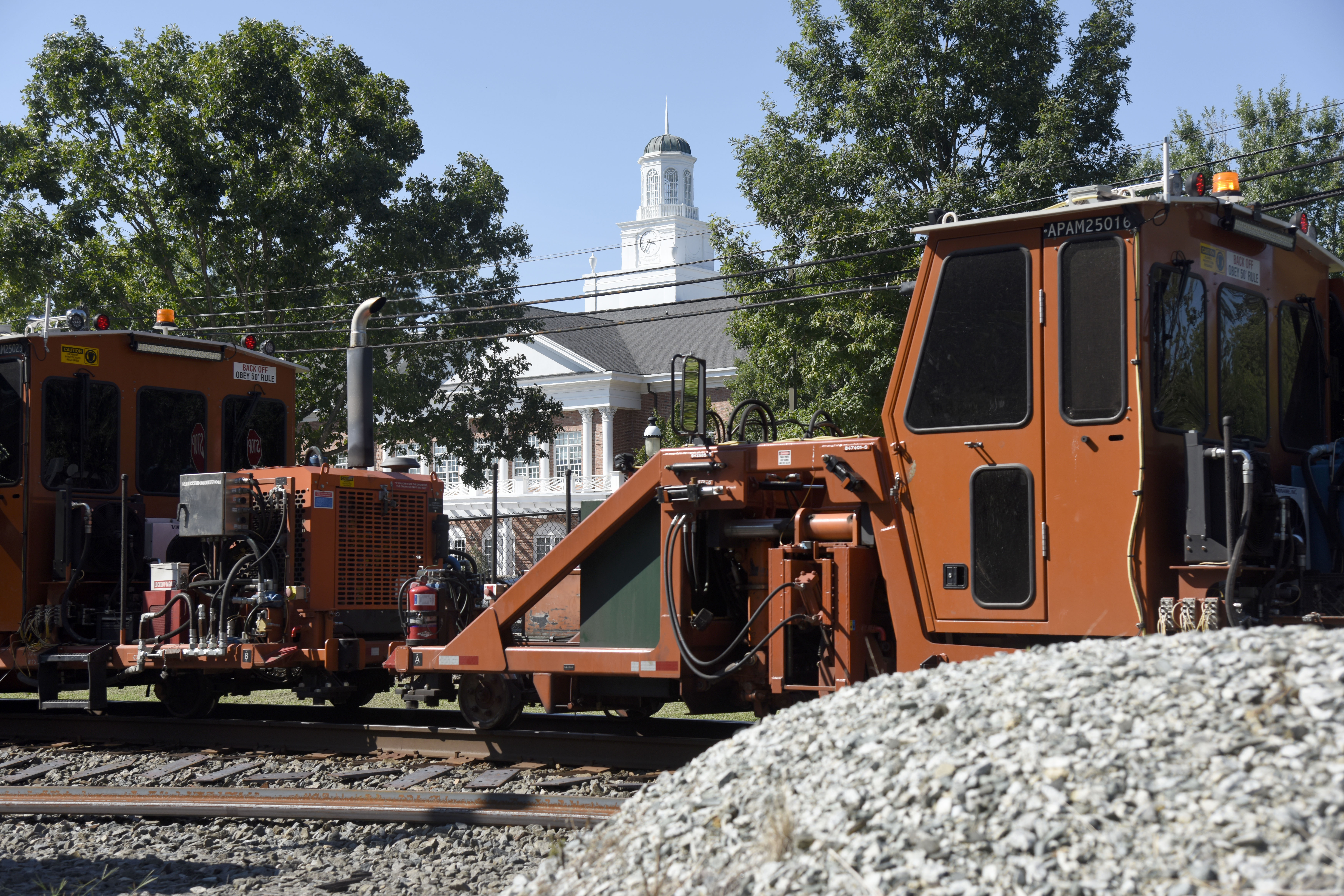 A photo of an orange Norfolk Southern rail maintenance piece of equipment on tracks near �������޹�����Ʒ with Lindner Hall in the background and a pile of railroad ballast in the foreground.