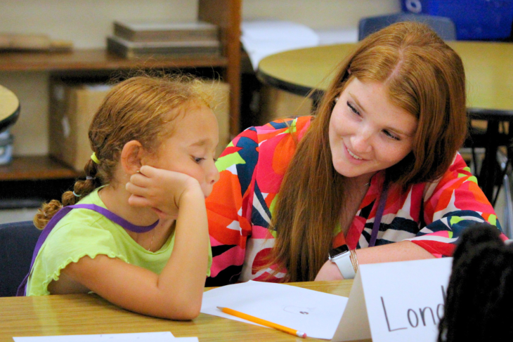 A teacher smiles while working one-on-one with a student at a classroom table.