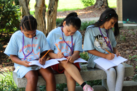 Three young girls sit on a bench in matching blue shirts and write in notebooks