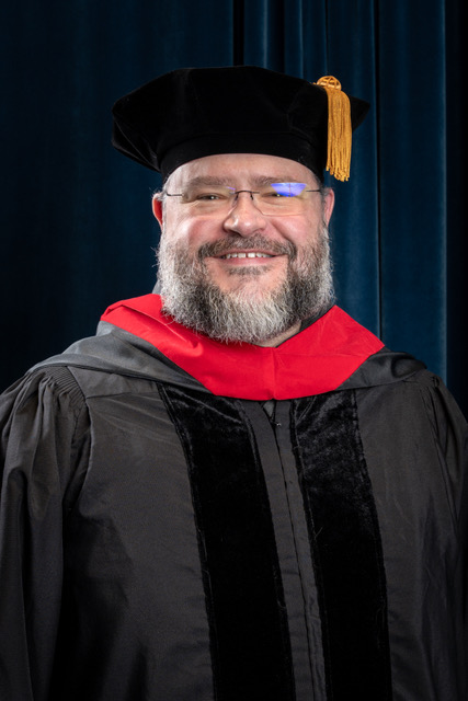 A smiling faculty member wearing black doctoral regalia, a red hood and a black velvet tam with a gold tassel poses for a formal portrait against a dark blue backdrop.