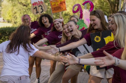 A student reaches out to greet cheering Elon students in maroon shirts holding colorful signs along a campus walkway.