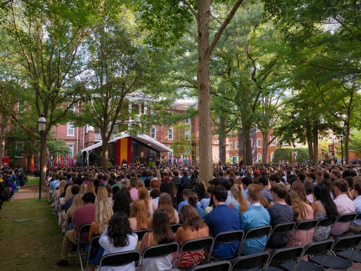 A photo of hundreds of students from the Class of 2029 watching speakers on a stage for New Student Convocation in August 2025. Large trees overhead provide shade.