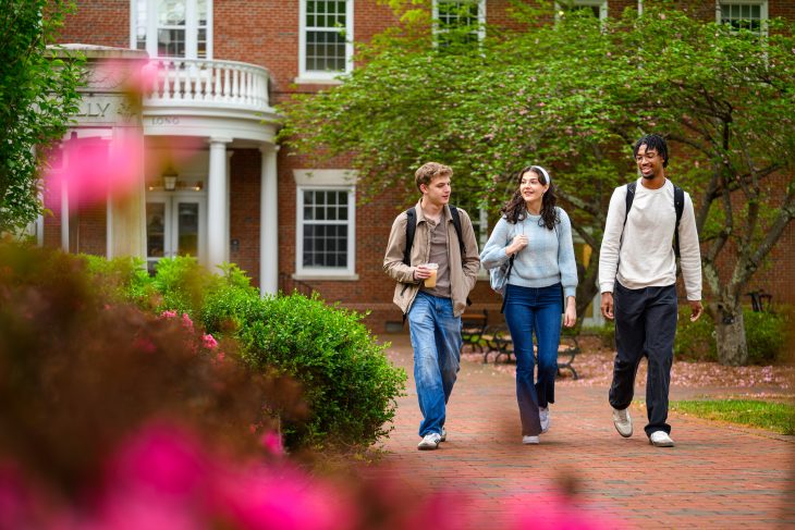 Three students carrying backpacks walk and talk together along a brick path surrounded by flowering trees and shrubs on Elon’s campus.