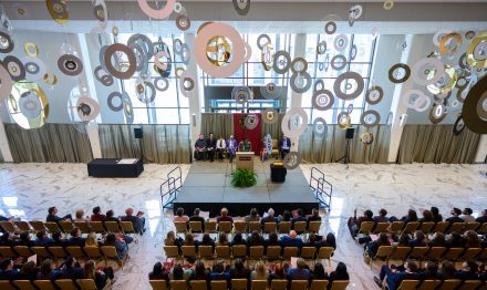 An overhead shot of a woman speaking at a podium on a stage to a large group of students seated in a theater atrium.